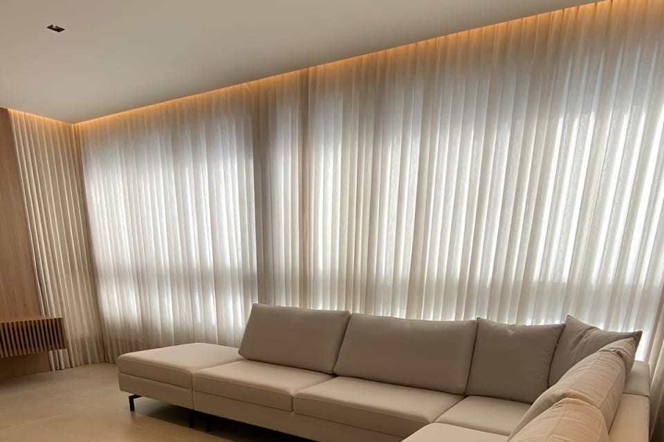 Sala ampla com cortina de voil em parede inteira e sofá bege, em Goiânia
